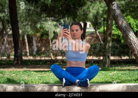 Eine ziemlich sportliche Frau, die im Park mit gekreuzten Beinen sitzt und ein Selfie auf ihrem Handy gemacht hat. Stockfoto