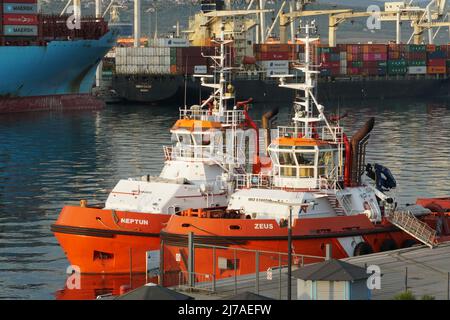 Zwei Schlepper mit orangefarbenem Rumpf, die im Hafen von Koper, Slowenien, vertäut sind. Im Hintergrund sind zwei Containerschiffe im Frachtbetrieb mit Portalkranen zu sehen. Stockfoto