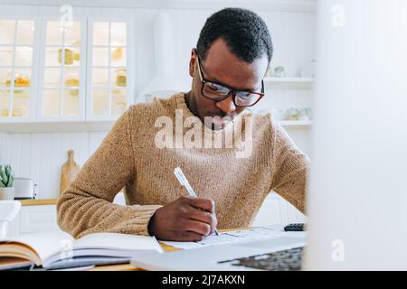 Der junge afroamerikanische Mann arbeitet online in einem Laptop, der Notizen am Tisch schreibt. Stockfoto