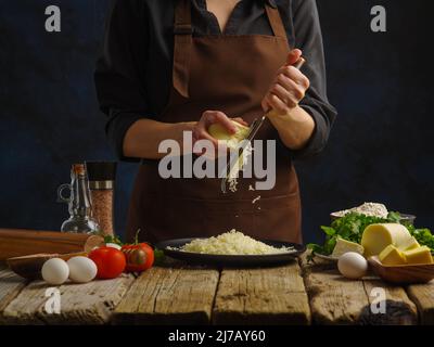 Ein professioneller Koch in dunkler Uniform reibt Käse. Gemüse, Kräuter, Eier, Gewürze - Zutaten für Salat, Pizza, Pasta. Gesunde vegetarische Kost, h Stockfoto