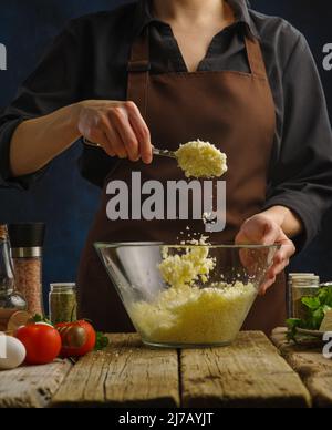 Der Küchenchef mischt geriebenen Käse in einer Glasschüssel. Kochen von Gerichten mit Spaghetti-Käse, Kuchen, Pizza, Pasta. Dunkelblauer Hintergrund. Nahaufnahme. Kochbuch, Essen blo Stockfoto