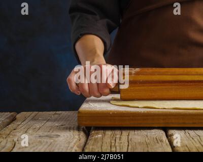 Kochen von Teigprodukten durch die Hände eines professionellen Küchenchefs auf einem hölzernen Küchentisch. Der Koch rollt den Teig auf einem Schneidebrett mit einem rollenden p aus Stockfoto