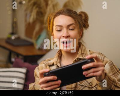 Ein Mädchen spielt ein Videospiel auf einer tragbaren Spielkonsole. Reagiert emotional auf das Spiel. Virtual Reality, Cyberspace, Cybersport, neue moderne Technologien Stockfoto