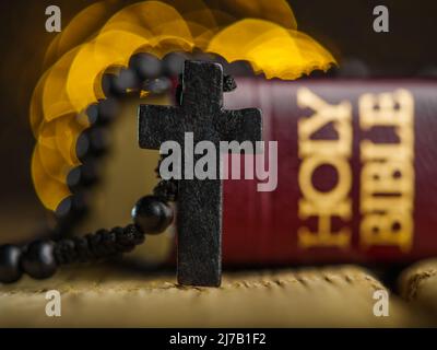 Symbole der Spiritualität, des Katholizismus - die Heilige Bibel und ein Kruzifix auf einem Rosenkranz auf einem Holztisch. Der Hintergrund ist gelb flackernde Lichter. Holy S Stockfoto