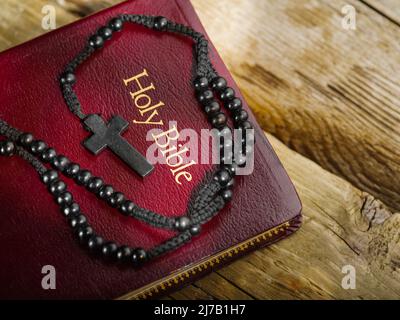 Auf einem einfachen Holztisch liegt ein Buch - die Heilige Bibel und ein Rosenkranz mit einem Kruzifix. Durch Sonnenlicht beleuchtet. Auf dem Foto befinden sich keine Personen. Da bin ich Stockfoto