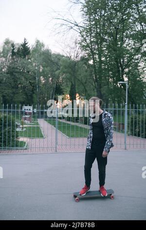 Lässiger Mann mit Longboard bei Sonnenuntergang. Hipster-Skater mit Skateboard. Junger Mann mit Schnurrbart und Bart-Langborading in der Parkstadt Stockfoto