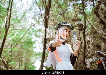 Schöne sportliche Frau in Hijab hält Flasche mit Daumen nach oben Stockfoto