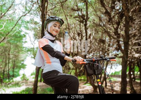 Attraktive sportliche Frau in Hijab hält Flasche beim Radfahren Stockfoto