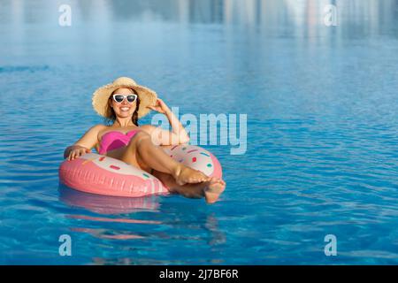 Lächelnde junge Frau in Strohhut und Sonnenbrille, die sich auf einem aufblasbaren Ring im Schwimmbad entspannt Stockfoto