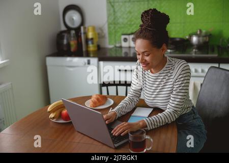 Junge positive Frau, die am Computer arbeitet, sitzt gut gelaunt am Tisch in der Küche Stockfoto