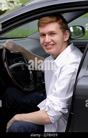 Schöner junger Mann mit riesigem Lächeln von seinem grauen Auto Stockfoto