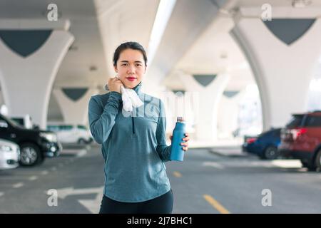 Asiatische Frau, die beim Joggen in der Stadt auf dem Parkplatz im Stadtgebiet ausruhte. Laufen und gesunde Outdoor-Zeit. Fit weibliche Person, die sportliche Aktivität