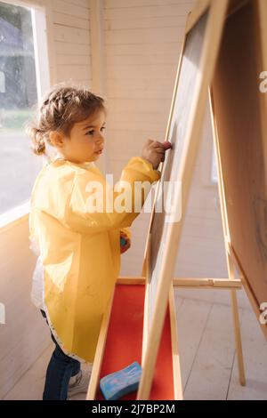 Der junge Künstler malt mit Kreide auf Holzbretter. Nettes Mädchen in yeallow Regenmantel Stockfoto