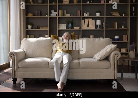 Entspannte, fröhliche Frau mittleren Alters, die auf einem bequemen Sofa sitzt Stockfoto