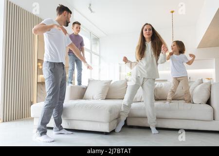 Lustige aktive Familie aus vier jungen erwachsenen Eltern und niedlichen kleinen Kindern, die im Wohnzimmer zusammen tanzen, sorglose kleine Kinder mit Mama Papa ha Stockfoto