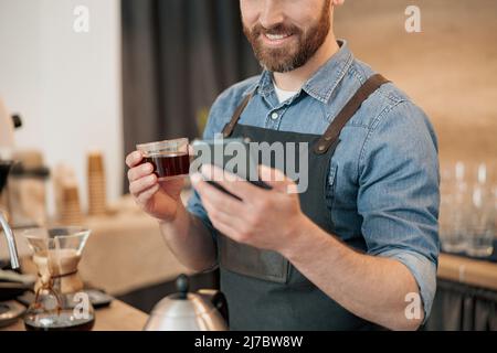Nahaufnahme des Barista, der während des Mittagessens Kaffee trinkt und durch die Social-Media-Feeds scrollt Stockfoto