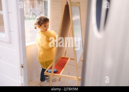 Der junge Künstler malt mit Kreide auf Holzbretter. Nettes Mädchen in yeallow Regenmantel Stockfoto