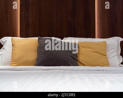 Set mit sauberen Kissen, braun, gelb und weiß, Decke und Bettlaken im Hotelzimmer mit dunklem Kopfteil aus Holz und Licht dahinter. Bettwäsche im Stockfoto