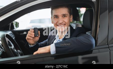 Glücklicher Versicherungsagent, der im neuen Auto sitzt, den Schlüssel vom Autofenster zeigt und die Kamera im Autohaus anlächelt Stockfoto