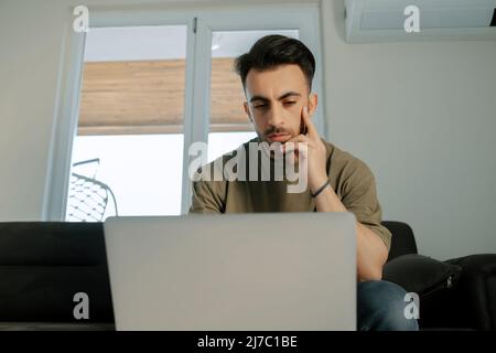 Nachdenklicher junger Mann, gutaussehender Mann, arbeitet online von zu Hause aus und versucht, ihr Geschäft aus der Ferne zu führen Stockfoto