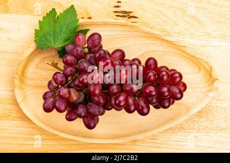 Draufsicht (Flat Lay) ein Bund reifer roter Trauben mit grünem Blatt in Holztablett auf Holztisch. Abunch von Crimson Seedless Trauben in einem Holztablett Stockfoto