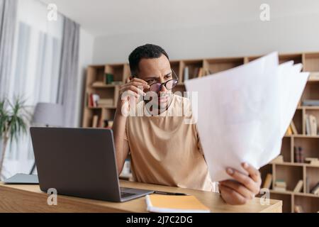 Ernsthafter junger arabischer Mann, der von zu Hause aus Dokumente liest und am Laptop arbeitet, sitzt am Schreibtisch Stockfoto