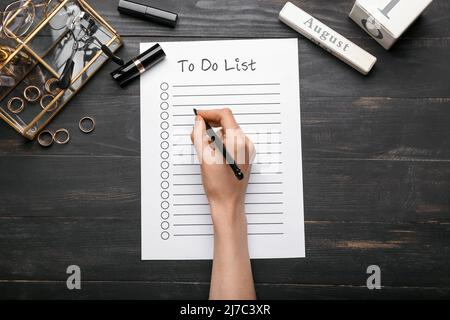 Weibliche Hand mit To-Do-Liste, Kosmetik und Zubehör auf dunklem Holzhintergrund Stockfoto