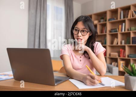 Fernschulungskonzept. Porträt einer fokussierten koreanischen Dame, die am Schreibtisch sitzt, einen Laptop benutzt und in einem Notebook schreibt Stockfoto