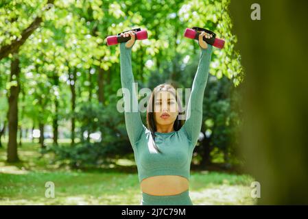 Eine attraktive junge Brünette Mädchen in einem olivgrünen Sport-Outfit heben Fitness Gewichte während der Outdoor-Workout-Übung im Stadtpark Stockfoto