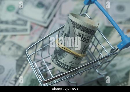 In einen Schlauch gerollte Dollars liegen in einem Einkaufskorb. Der Korb steht auf einem verschwommenen Geldhintergrund. Stockfoto