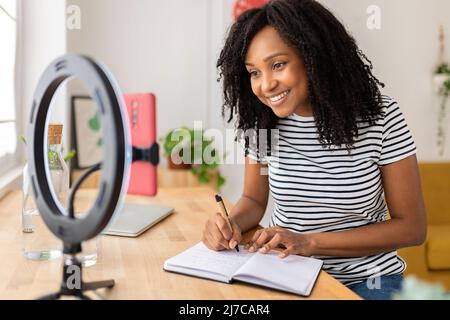 Junge afroamerikanische Frau, die sich Notizen gemacht hat, während sie sich Videos auf dem Mobiltelefon ansieht Stockfoto