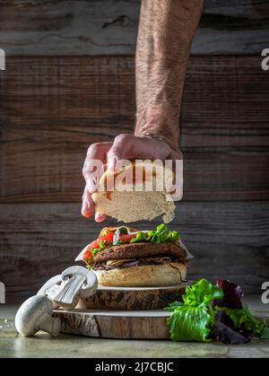 Hamburger mit Kichererbsen-Patty, Tomaten, Pilzen und Gemüse auf Roggenbrot. Eine Hand beendet die Zusammensetzung des Burgers.Copy Space.Vertikale Aufnahme Stockfoto