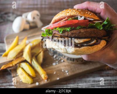 Hamburger mit Kichererbsen-Patty, Tomaten, Pilzen und Gemüse auf Roggenbrot. Eine Hand hält den Burger. Auf dem Boden Pommes und Pilze. Speicherplatz kopieren. Stockfoto