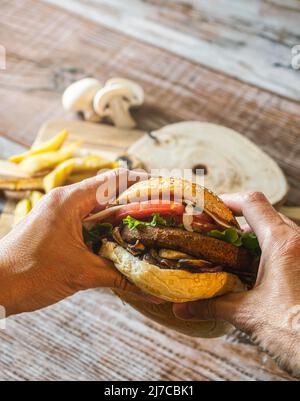 Hamburger mit Kichererbsen-Patty, Tomaten, Pilzen und Gemüse auf Roggenbrot. Zwei Hände halten den Burger. Auf dem Boden Pommes und Pilze.Platz kopieren. Stockfoto