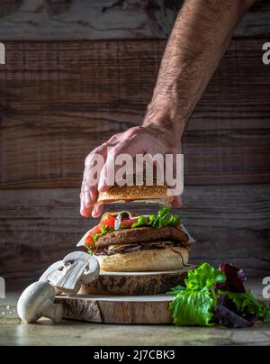 Hamburger mit Kichererbsen-Patty, Tomaten, Pilzen und Gemüse auf Roggenbrot. Eine Hand beendet die Zusammensetzung des Burgers.Copy Space.Vertikale Aufnahme Stockfoto