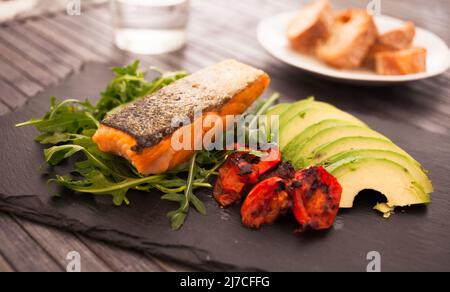 Gebackenes Meerforellenfilet mit frischem Gemüse und Kräutern auf schwarzem Brett Stockfoto