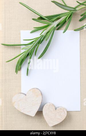 Rosmarin (Rosmarinus officinalis) Zweig, weiße Papierkarte und zwei hölzerne Herzen. Grafikressourcen Stockfoto