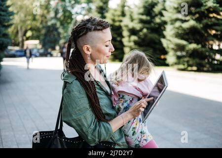 Freiberufliche Eltern. Beschäftigt arbeiten Mutter mit Baby Kleinkind Tochter auf die Hände gehen und suchen auf Tablet auf der Straße. Multitasking Freelancer Mama macht Notizen Stockfoto