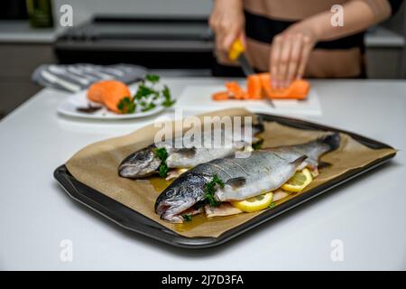 Kaukasische Frau in der Küche, die die Zutaten für ein gesundes Abendessen zubereitet. Sie stellte Regenbogenforelle mit Zitronenscheiben auf ein Backblech in einer Bratpfanne Stockfoto