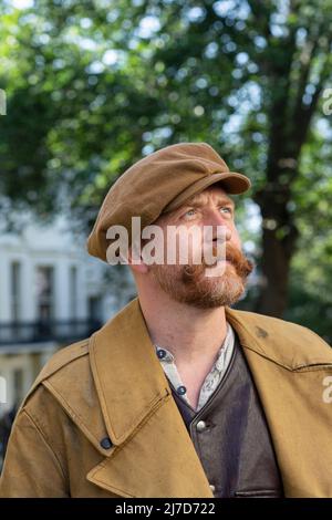 Ein Mann in einer flachen Mütze im altmodischen Outfit auf der Straße mit Baum im Hintergrund. Stockfoto