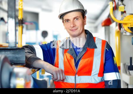 Junger positiver Ingenieur in Helm und Konstruktionsweste blickt in die Kamera und lächelt. Porträt eines jungen Schwerarbeiters im Raum mit unscharfem Hintergrund. Mitarbeiter in der Gasindustrie. Stockfoto