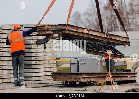 Slinger in Helm und Weste steuert das Entladen von Metallkonstruktionen auf der Baustelle. Weißer Handwerker entlädt Ladung. Stockfoto