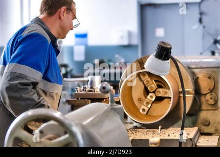 Turner in Overalls arbeitet hinter der Drehbank in der Produktionshalle. Verarbeitung von Metallprodukten. Industrieller Hintergrund. Stockfoto