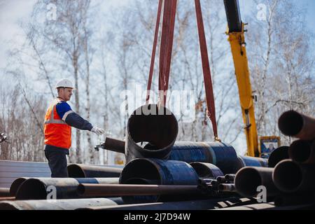 Slinger im Helm entlädt am sonnigen Sommertag Metallrohre auf der Baustelle. Echte Szene. Workflow. Echter Arbeiter. Stockfoto