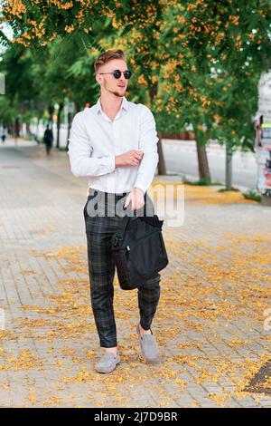 Geschäftsmann, der eine Sonnenbrille trägt und eine Tasche in der Hand hält, während er draußen auf der Straße läuft. Stockfoto