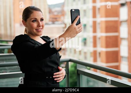 Junge Geschäftsfrau, die mit einem Mobiltelefon ein Selfie gemacht hat, während sie draußen auf einem Balkon steht. Stockfoto