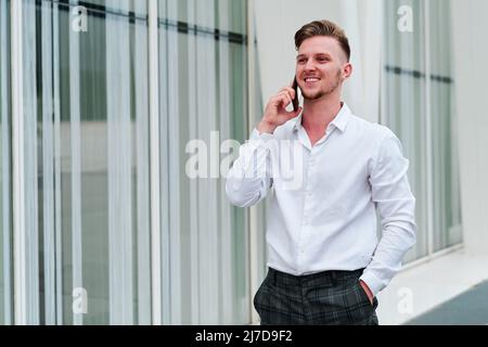 Ein junger Geschäftsmann lächelt, während er vor dem Bürogebäude telefoniert. Stockfoto