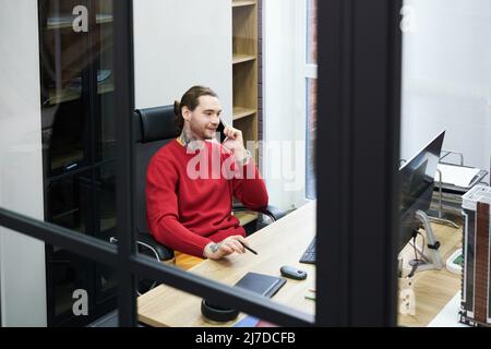 Lächelnder Manager sitzt am Tisch im Büro vor dem Computermonitor und führt Geschäftsgespräche auf dem Mobiltelefon Stockfoto