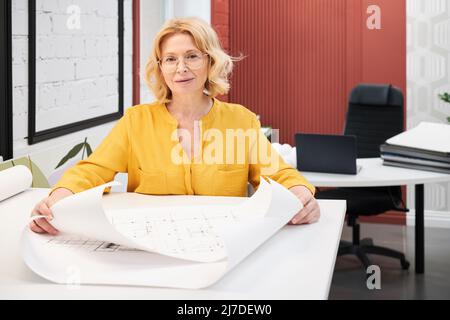 Porträt einer reifen Architektin in Brillen, die die Kamera anschaut, während sie mit Blaupausen von neuen Gebäuden am Tisch arbeitet Stockfoto