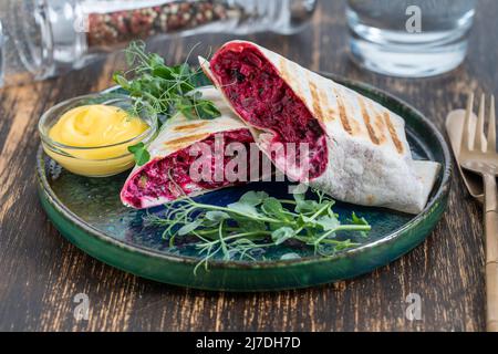 Veganer Burrito. Aufgeschnittene rohe Lebensmittelverpackung mit roter Rote Beete, Feta-Käse, Pflaumen, Dill und Rahmsoße mit Erbsen-Mikrogemüse auf einem Teller. Nahaufnahme Stockfoto